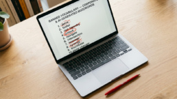 A high-angle, editorial photograph of a laptop on a light wood desk. The laptop screen displays a document titled 'Banned Vocabulary — Corporate & AI-Generated Buzzwords' with a numbered list including words like 'delve,' 'rich tapestry,' and 'synergy.' Several words are aggressively circled or crossed out with red ink. A red pen sits on the desk next to the laptop, and the scene is illuminated by soft, natural light.